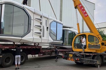 Crane and truck handling space capsule cabins beside a factory building